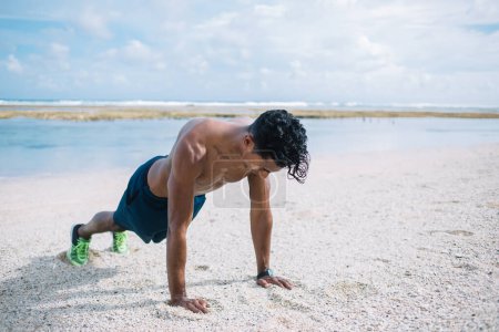 Strong sportive bare chested Hispanic male wearing shorts and sneakers doing push up exercise while working out on sandy beach