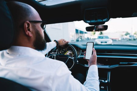 Photo for Back view of executive driver using cellphone technology for tracking gps location via smartphone application while steering to destination, businessman doing automotive wheeling and phoning - Royalty Free Image