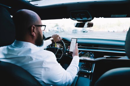 Photo for Back view of male driver reading received email message while steering automotive vehicle, employer in white shirt checking mobile time via cell application while lateness to business meeting - Royalty Free Image