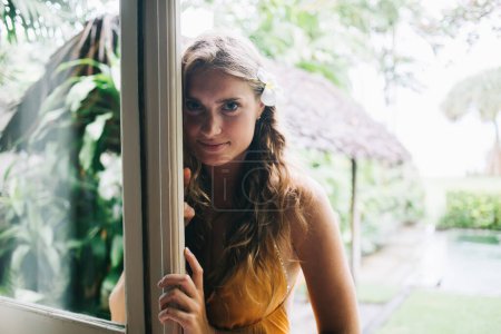 Attraktive, sanfte, erwachsene blonde Frau in orangefarbenem Kleid mit Orchidee im Haar, die Kamera schaut und lächelt, während sie an der Tür steht und auf verschwommenem Hintergrund des tropischen Gartens im Bali Resort bei sonnigem Tag lehnt