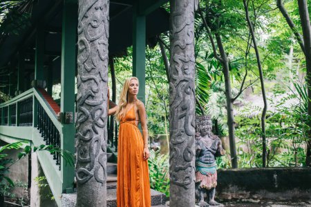 Attraktive modische Frau mit blondem Haar lehnt an Steinsäule mit Stuckverzierungen buddhistischer Statue und blickt auf Bali weg