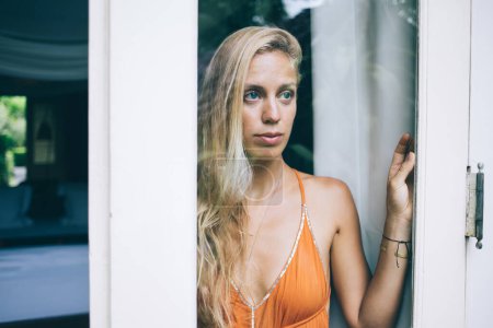 Téléchargez les photos : Vue à travers le verre de réfléchi jeune femme blonde charmante rêvant de la vie et regardant loin tout en se tenant à la porte à l'intérieur de la maison - en image libre de droit