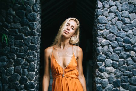 Attractive relaxed slim blonde female wearing orange dress with deep neckline standing with head tilted to side between dark grey stone walls