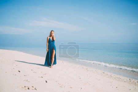 Blonde young calm female in long blue dress touching hair and walking in relaxed manner along seashore while looking at camera