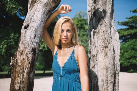 Young calm female in elegant blue dress standing between trees at sand beach and touching tree while looking at camera 