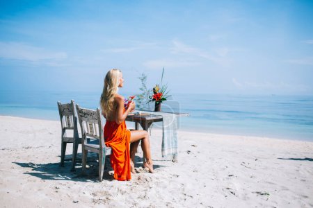 Contenido sonriente dama feliz en vestido naranja soñando mientras se mira en la distancia del océano durante la bebida en la playa idílica exótica soleada
