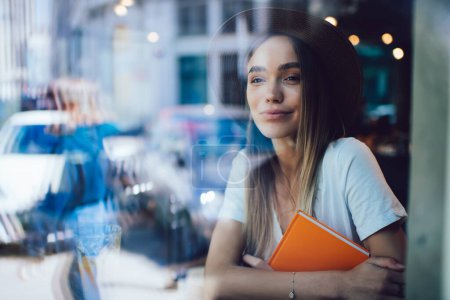 Téléchargez les photos : Superbe femme insouciante en chapeau et T-shirt tendance assis dans un café et regardant loin tout en embrassant le livre orange et en contemplant la rue - en image libre de droit