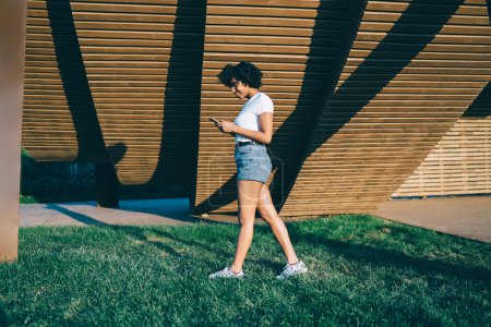 Foto de Atractiva chica hipster de piel oscura en pantalones vaqueros caminando charlando en línea a través de teléfono inteligente recreando en el día soleado, feliz afroamericano chica hipster millennial comprobar correo electrónico en el teléfono móvil - Imagen libre de derechos