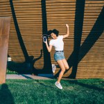 Joyful glad ethnic lady with curly hair dressed in white shirt and shorts dancing listening to music with earphones in street in summer 