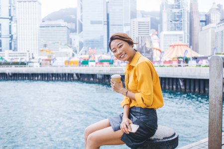 Portrait of happy female tourist with tasty caffeine beverage taking rest on city area with Hong Kong on background, cheerful Japanese woman with coffee to go recreating during Chinese vacations