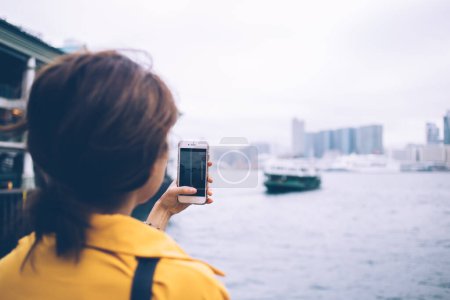 Foto de Vista trasera de una turista irreconocible usando una blusa amarilla tomando una foto vertical de un ferry verde y rascacielos modernos en la bahía de la ciudad de Hong Kong en un teléfono celular blanco - Imagen libre de derechos