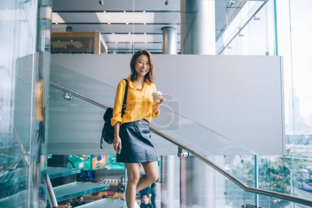 Young happy cheerful Asian female in yellow shirt black leather skirt and backpack going down staircase with hot drink and cellphone in Hong Kong