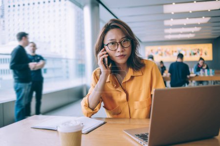 Téléchargez les photos : Asiatique attrayant étudiant féminin en chemise jaune et lunettes appelant au bureau avec ordinateur portable et café et regardant la caméra dans la salle à manger à l'université de Hong Kong - en image libre de droit