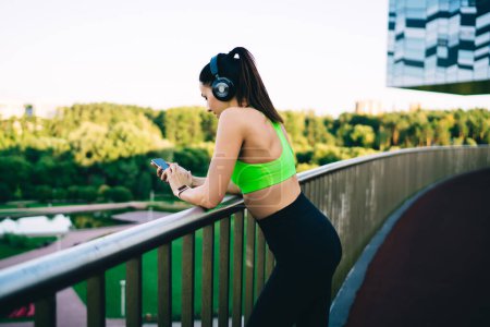 Rear view of young caucasian woman jogger in headphones using smartphone for checking app notifications on workout break, sporty female in active wear share media via smartphone in blog about fitness