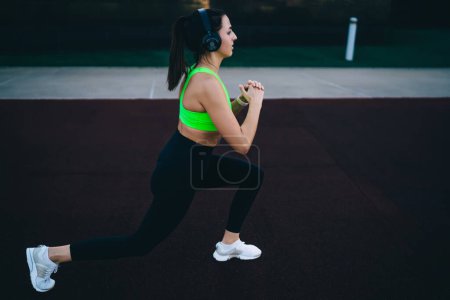 Side view of strong female athlete in active wear and headphones training legs and ass muscles outdoors, sportswoman listening vitality playlist via electronic accessory having morning ABT workout
