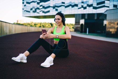 Joyeux caucasien femme jogger lecture message sur téléphone mobile ayant pause sur l'entraînement, souriant 20s femme sportive dans les écouteurs télécharger de la musique sur smartphone pour l'entraînement actif à l'extérieur