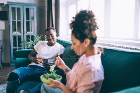 Young African American man in glasses and casual clothes drinking juice and using phone while woman having lunch sitting together on sofa in living room