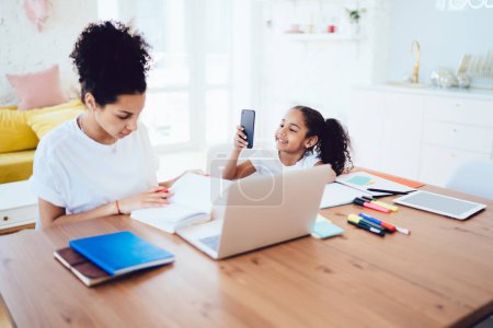 Photo for African American daughter taking photo of mother while working remotely sitting at wooden table with laptop spending time together at home - Royalty Free Image