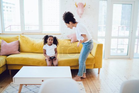 Photo for Angry African American woman standing in bright room and screaming at daughter while kid sitting on sofa with controller and looking at camera - Royalty Free Image