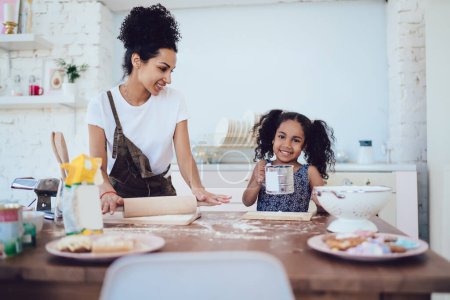Lächelndes fröhliches Kind mit Mehlsieb hilft fröhlicher emsiger Mama beim Kochen von Backwaren auf dem Küchentisch, während sie in die Kamera schaut