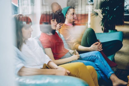 Durch Glas sitzen nachdenkliche junge Leute in bunten Freizeitklamotten auf einem weichen türkisfarbenen Sofa in der hellen, gemütlichen Lobby des modernen Gebäudes