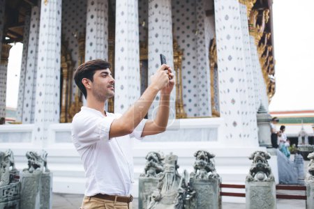 Photo for Positive spanish hipster guy using smartphone camera taking picture of asian architecture and temples during sightseeing tour, positive travel blogger shooting video for vlog in Thai on destinatio - Royalty Free Image