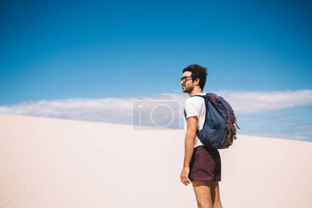 Jeune touriste sud-asiatique en lunettes de soleil branchées regardant autour des dunes sauvages dans la vallée de la mort, vagabond indien avec sac à dos passer des vacances d'été pour visiter safari inhabité au Maroc
