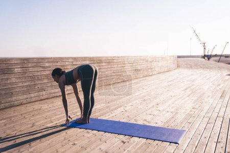 Foto de Vista lateral de una mujer atenta y bien concentrada en ropa deportiva acogedora contemporánea que practica yoga sobre una alfombra estrecha azul en pista a plena luz del día - Imagen libre de derechos