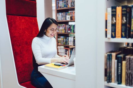 Photo for Smiling positive cheerful Asian woman typing on laptop during studying while sitting in individual study space in library among bookshelves - Royalty Free Image