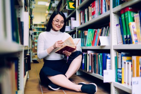 Photo for Smiling Asian woman sitting with crossed legs on floor among bookshelves flipping pages of book while searching for educational literature in library - Royalty Free Image