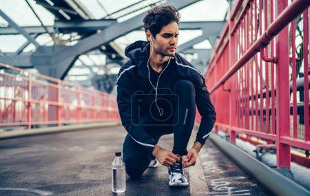 Active male jogger tying laces of stylish sneakers for start morning running at city urbanity, European crossfit bodybuilder in electronic headphones listening motivated audio book for warm up