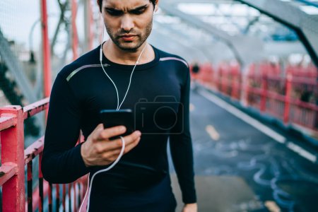 Millennial männlicher Sportblogger erstellt Veröffentlichung für eigene Website mittels App auf Smartphone in Laufpause, europäischer Sportler aktualisiert Playlist auf Handy vor dem Training im Freien