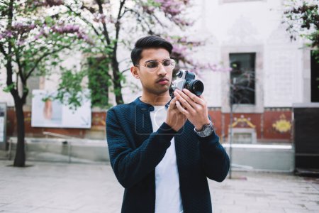 Serious hindu male tourist in trendy wear using camera for photographing during vacations on urban settings, pensive hipster guy holding equipment for taking picture of city having journey on weekends