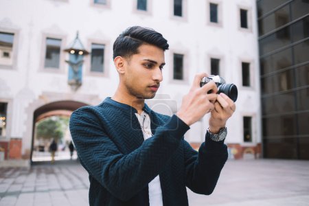 Serious hindu male traveler in trendy wear making photo of city using camera during vacation trip, pensive 20s man amateur spending time on hobby concentrated in taking picture in town square