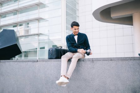 Cheerful hindu male traveler sitting on urban setting with bag and camera enjoying free time and vacation journey, smiling hipster guy checking equipment and photos on holiday in city during trip