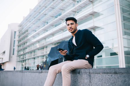 Portrait of handsome hindu male in stylish wear and spectacles using mobile phone for chatting and communication on urban setting, serious confident businessman sitting on city square on leisure