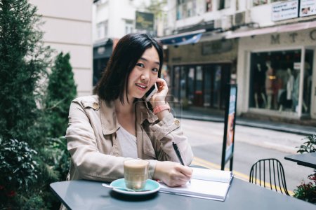 Foto de Mujer asiática joven desconcertada con el pelo negro corto sentado en el escritorio con una taza de café y hablando en el teléfono inteligente mientras escribe en el cuaderno contra el fondo de la carretera de la ciudad - Imagen libre de derechos