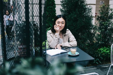 Foto de Joven mujer asiática alegre en ropa casual sentada en la mesa con cuaderno y sonriendo mientras mira a la cámara y toca la barbilla en la cafetería de la calle cerca de las plantas - Imagen libre de derechos