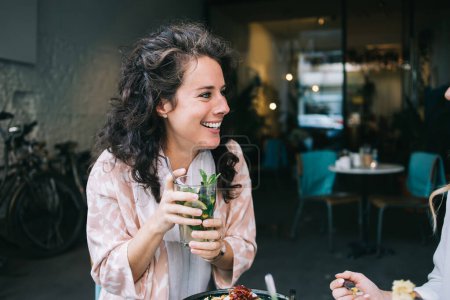 Téléchargez les photos : Joyeux caucasien femme en tenue tendance recréant sur le temps libre tenant cocktail frais avec de la glace et de la menthe, souriant 20s femme aux cheveux bouclés rire sur la pause déjeuner profiter de boisson mojito - en image libre de droit
