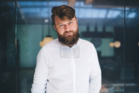 Half length portrait of masculine bearded businessman dressed in white smart casual shirt with copy space for brand name, confident Caucasian male entrepreneur 30s posing in modern workspace