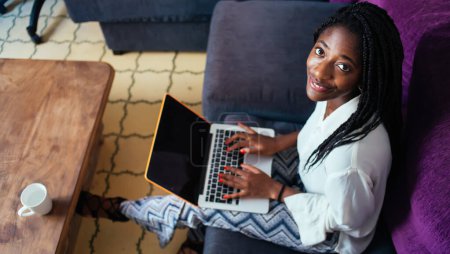 Téléchargez les photos : Vue du dessus de la jeune femme à la peau foncée freelance travaillant à distance de la maison assis à un canapé confortable, souriant femme afro-américaine en utilisant un ordinateur portable pour la réservation et effectuer des paiements - en image libre de droit