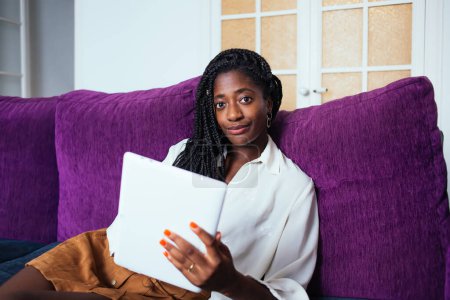 Téléchargez les photos : Portrait de femme à la peau foncée recréant sur un canapé confortable dans le salon à l'aide d'une tablette numérique moderne pour bavarder, attrayante blogueuse afro-américaine sur les réseaux sociaux via pavé tactile à la maison - en image libre de droit