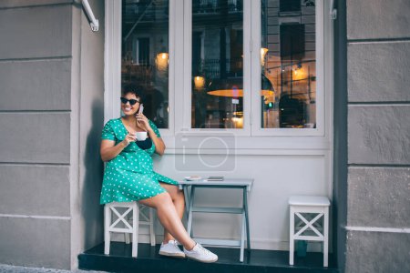 Spanierin mit trendiger Sonnenbrille genießt freundliche Smartphone-Gespräche während der Kaffeezeit in der Cafeteria auf dem Bürgersteig, fröhliches Plus-Size-Modell telefoniert per Handy-Applikation mit drahtlosem Anschluss