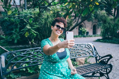 Glückliche Frau mit trendiger Sonnenbrille, die für Selfie-Klicks posiert und soziale Netzwerke nutzt, um Reiseblogs zu popularisieren, fröhliche spanische Influencerin, die vor der Smartphone-Kamera lächelt