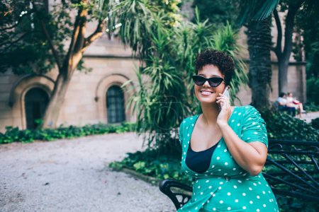 Lächelnder hispanischer Tourist, der sich auf einer Bank ausruht, um ein internationales Handy-Gespräch mit einem Freund zu führen und über den Laubpark zu sprechen, Porträt einer zahmen Reisenden im gepunkteten Kleid, die per Handy anruft