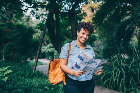 Halblanges Porträt einer fröhlichen Touristin mit Rucksack und Papierkarte, die ihren Urlaub genießt, um die Welt zu erkunden, fröhliches hispanisches Mädchen mit Positionspapier, das im Stadtpark in die Kamera lächelt