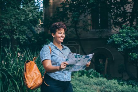 Fröhlich lächelnde junge ethnische Frau in Freizeitkleidung mit Rucksack, der Papierkarte liest und im Park spazieren geht und wegschaut