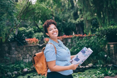 Téléchargez les photos : Voyageur ethnique paisible en tenue décontractée avec sac à dos et carte papier explorant la ville se promener dans un parc tropical en regardant la caméra - en image libre de droit