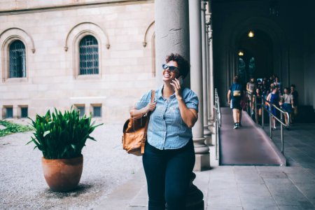 Fröhliche Frau in lässiger Kleidung und trendiger Sonnenbrille beim freundlichen Handy-Gespräch lachend, glückliche Studentin mit Rucksack-Anruf per Handy in der Uni-Pause