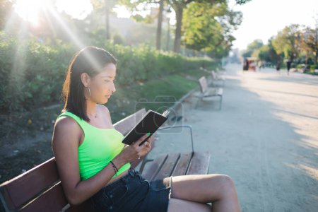 Thoughtful brunette female student sitting on bench with book concentrated on learning information outdoors, pensive 20s trendy dressed hipster girl enjoying reading hobby in park on sunny day
